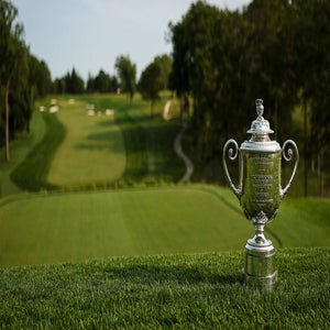 A silver golf trophy sits on lush grass in the foreground, celebrating PGA Championship glory, with a manicured golf course and trees stretching out in the background under a clear sky.