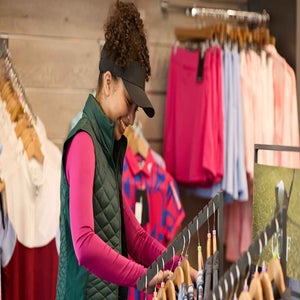 A woman wearing a visor, pink long-sleeve shirt, and green vest smiles while shopping for clothes in a store, browsing a rack of colorful shirts&mdash;perfect for finding a thoughtful Mother&rsquo;s Day gift.