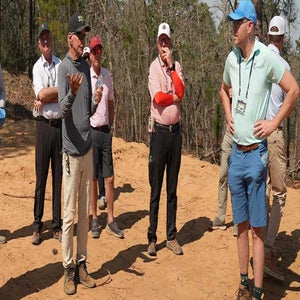 A group of men stand outdoors on sandy ground, some wearing hats and sunglasses, discussing golf architecture. One man in front gestures with his hands while others listen. Trees and sparse vegetation are visible in the background.
