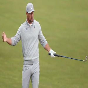 A golfer, reminiscent of Bryson DeChambeau, wearing a gray cap and sweater, gestures with one hand while holding a golf club in the other, standing on a grassy PGA Championship golf course.