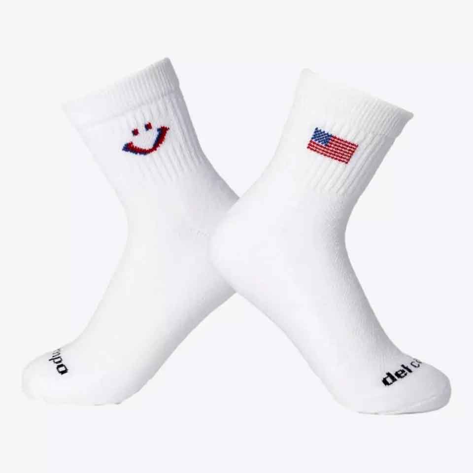 Two white socks crossed at the ankle—one with a small American flag design, the other with a smiley face made from red and blue stitches. Both feature black text near the toes, making them a fun Father's Day gift.