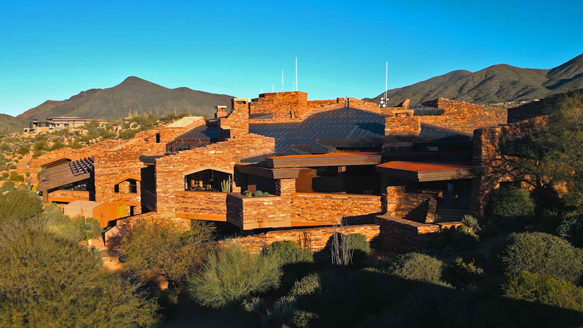 A sprawling building made of reddish stone blends into a desert mountain landscape, surrounded by shrubs and mountains under a clear blue sky.