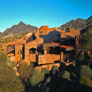 A sprawling building made of reddish stone blends into a desert mountain landscape, surrounded by shrubs and mountains under a clear blue sky.