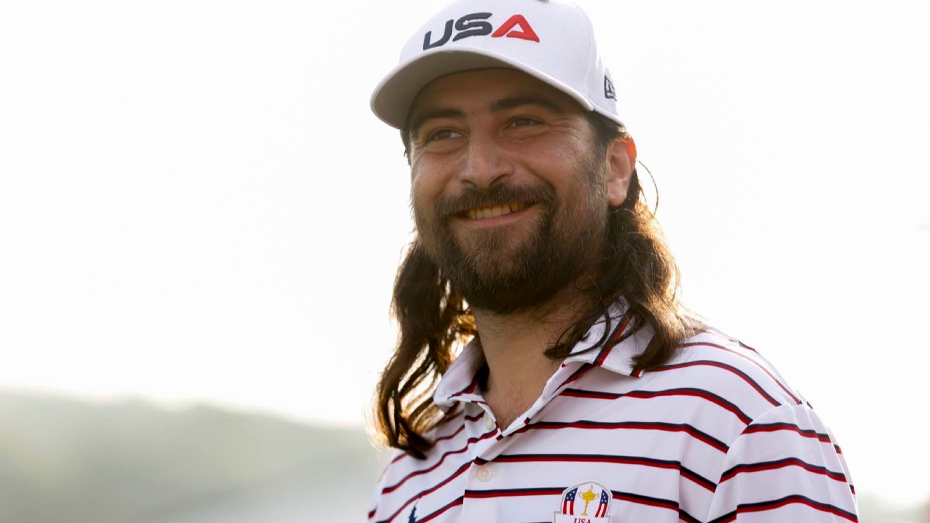 Noah Kahan with long brown hair and a beard wears a white USA cap and a white, red-striped shirt with an embroidered Ryder Cup logo, standing outdoors in soft sunlight, as if ready to share some Noah Kahan golf advice.