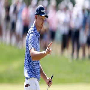 A golfer in a blue striped shirt and cap holds a putter and gestures with two fingers raised, while standing on a golf course with a blurred crowd in the background.