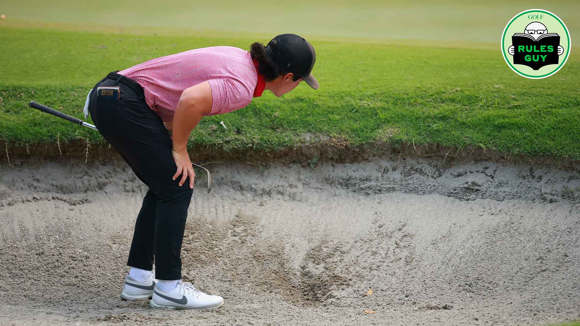 A golfer in a red shirt and black pants bends over to examine an embedded ball in a sand bunker, with a golf club resting on their back and the “Rules Guy” logo in the top right corner. A grassy green is visible in the background.