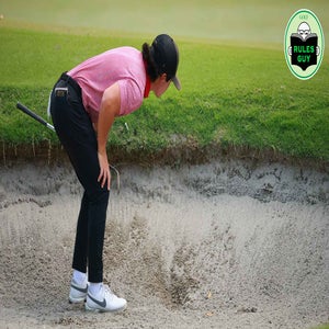 A golfer in a red shirt and black pants bends over to examine an embedded ball in a sand bunker, with a golf club resting on their back and the &ldquo;Rules Guy&rdquo; logo in the top right corner. A grassy green is visible in the background.