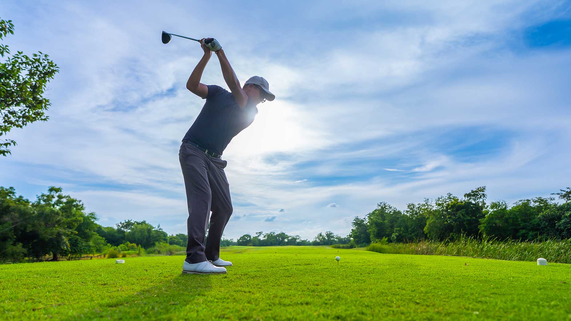 A golfer in a black shirt, gray pants, and white shoes prepares to swing a club during a range session on a bright, sunny golf course, with blue sky and trees in the background.