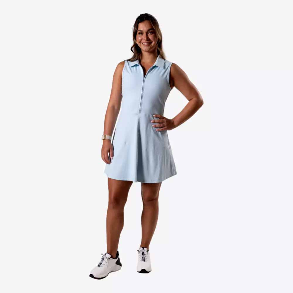 A woman stands smiling with one hand on her hip, dressed in elegant attire—a light blue sleeveless dress and white sneakers—against a plain white background, perfect for a Mother's Day look.