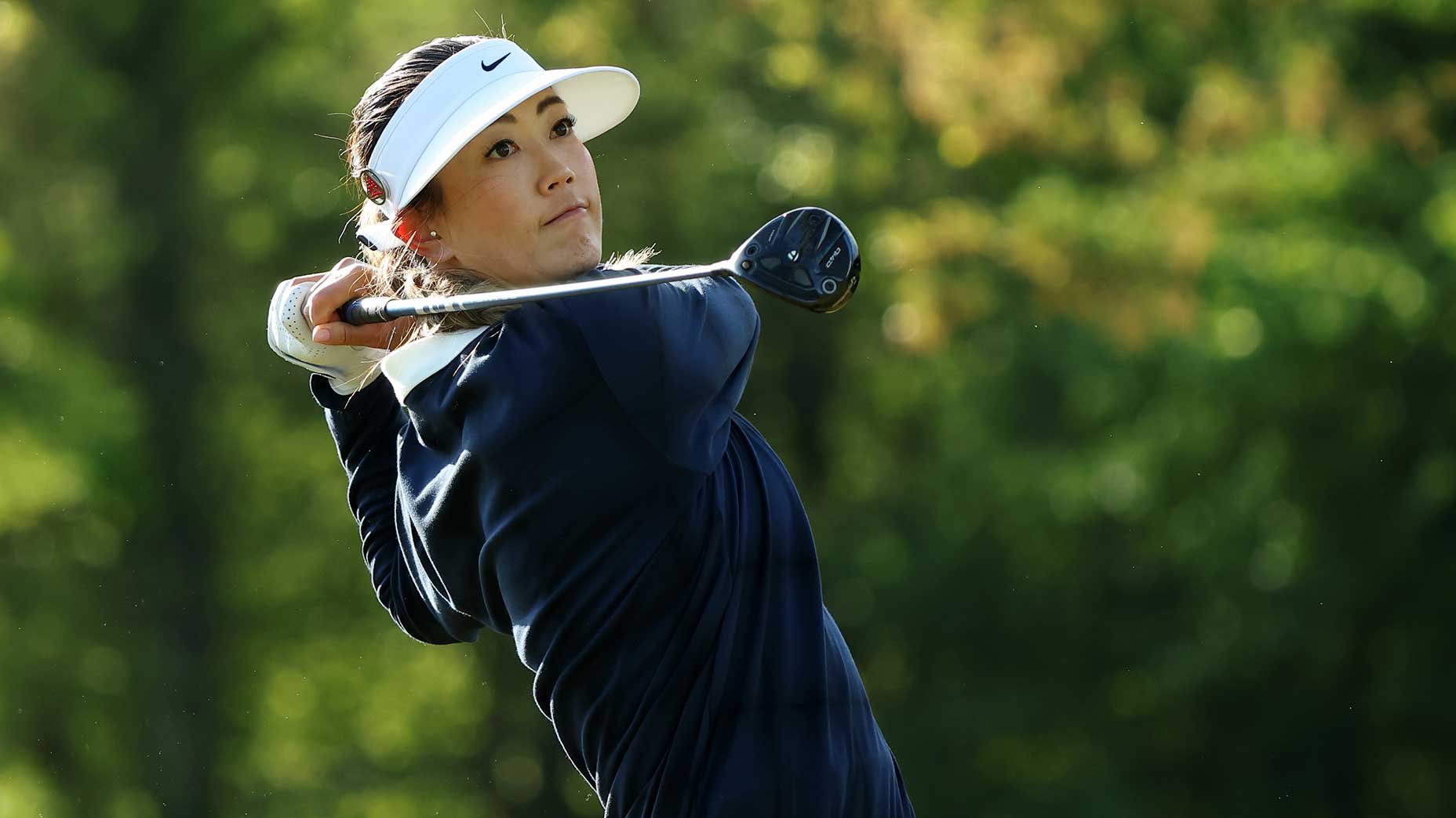 Michelle Wie West, wearing a white visor and navy jacket, swings a golf club outdoors, framed by a blurred green background of trees and sunlight.