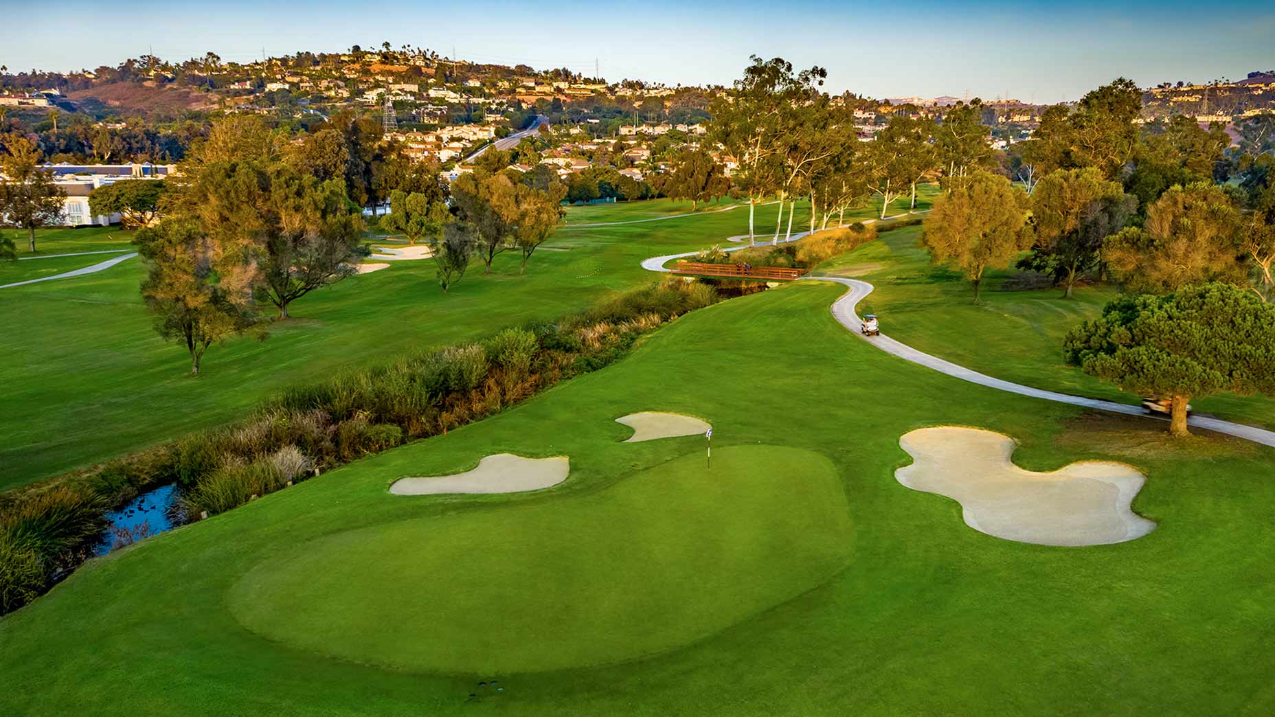Aerial view of the lush green Omni La Costa golf course, featuring sand bunkers, winding paths, trees, and a distant hillside neighborhood under a clear sky.