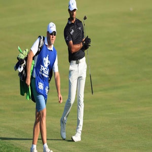 Aaron Rai in a black shirt and white pants stands on a fairway holding a club, while a caddie in a blue vest and white shirt walks ahead carrying a golf bag. Both are on a well-maintained grassy course.