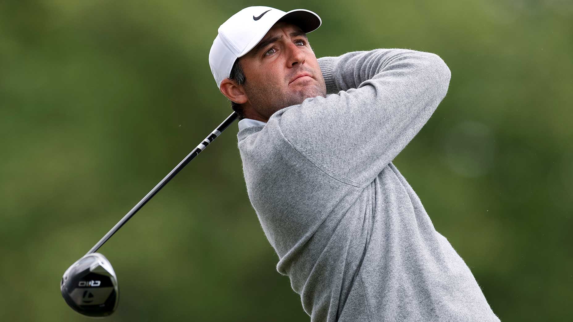 A golfer wearing a gray sweater and white cap swings a golf club, focusing intently on his shot. The background is blurred with green foliage, capturing the excitement of the 2026 PGA Championship live updates.