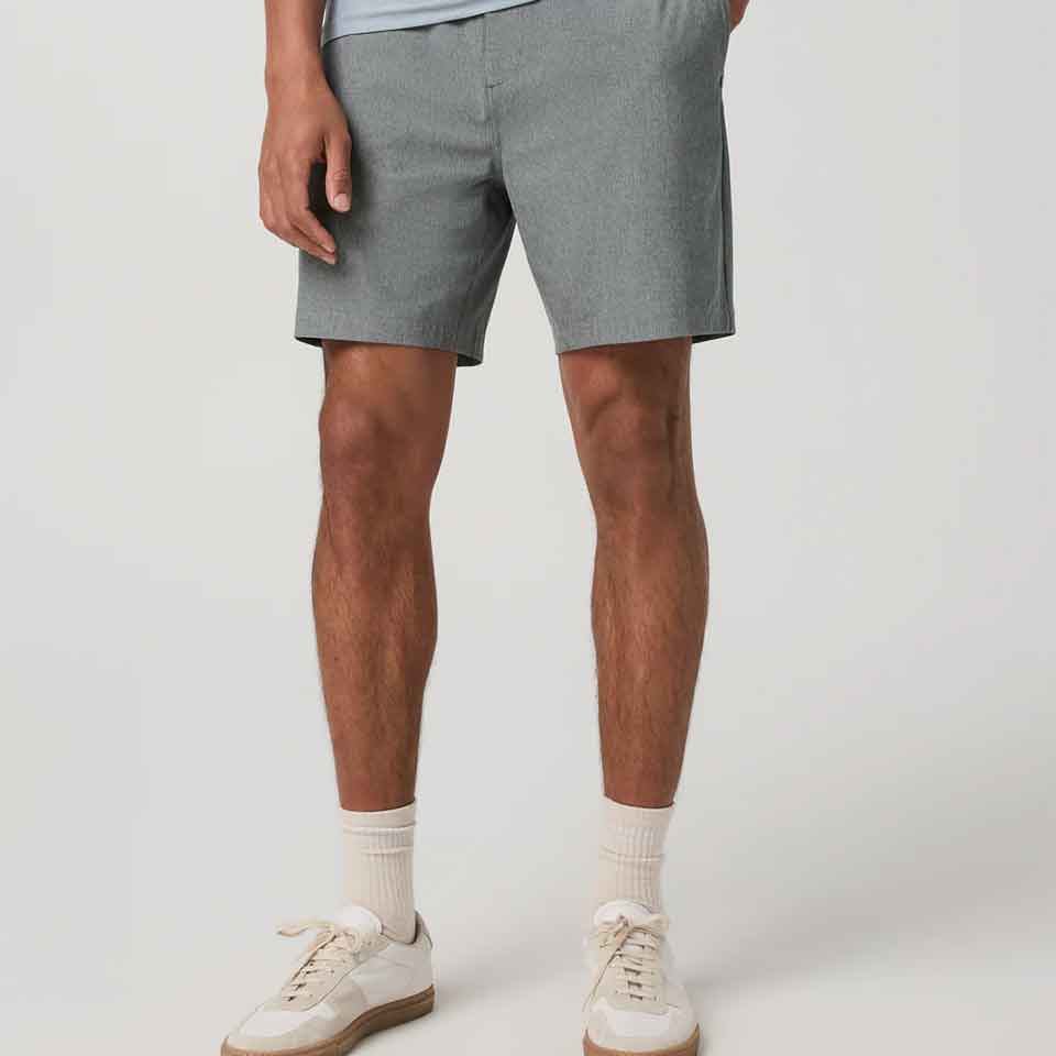 A person wearing light gray Vuori shorts, a pale blue shirt, white crew socks, and white sneakers, standing against a plain light background.