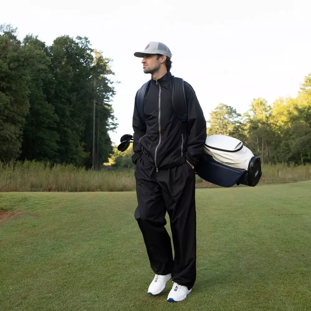 A person wearing black tracksuits, white sneakers and a gray cap stands on a grass field holding a golf bag – perhaps inspired by the 6 golf deals worth buying right now – with trees and blue sky in the background.