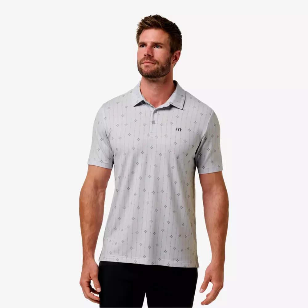 A man with short brown hair and a beard wears a light gray short-sleeved polo shirt with a subtle polka dot pattern and black pants, standing against a plain white background, ready to match his look with the best golf smart watch.