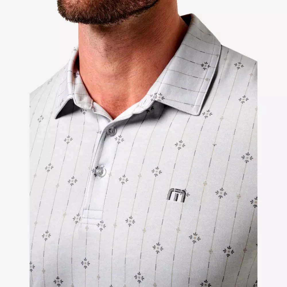 A close-up of a man wearing a light gray short-sleeved polo shirt with a subtle, dark geometric pattern and an embroidered logo on the chest. The shirt has a collar, two buttons, and he also has the best golf smart watch on his wrist.