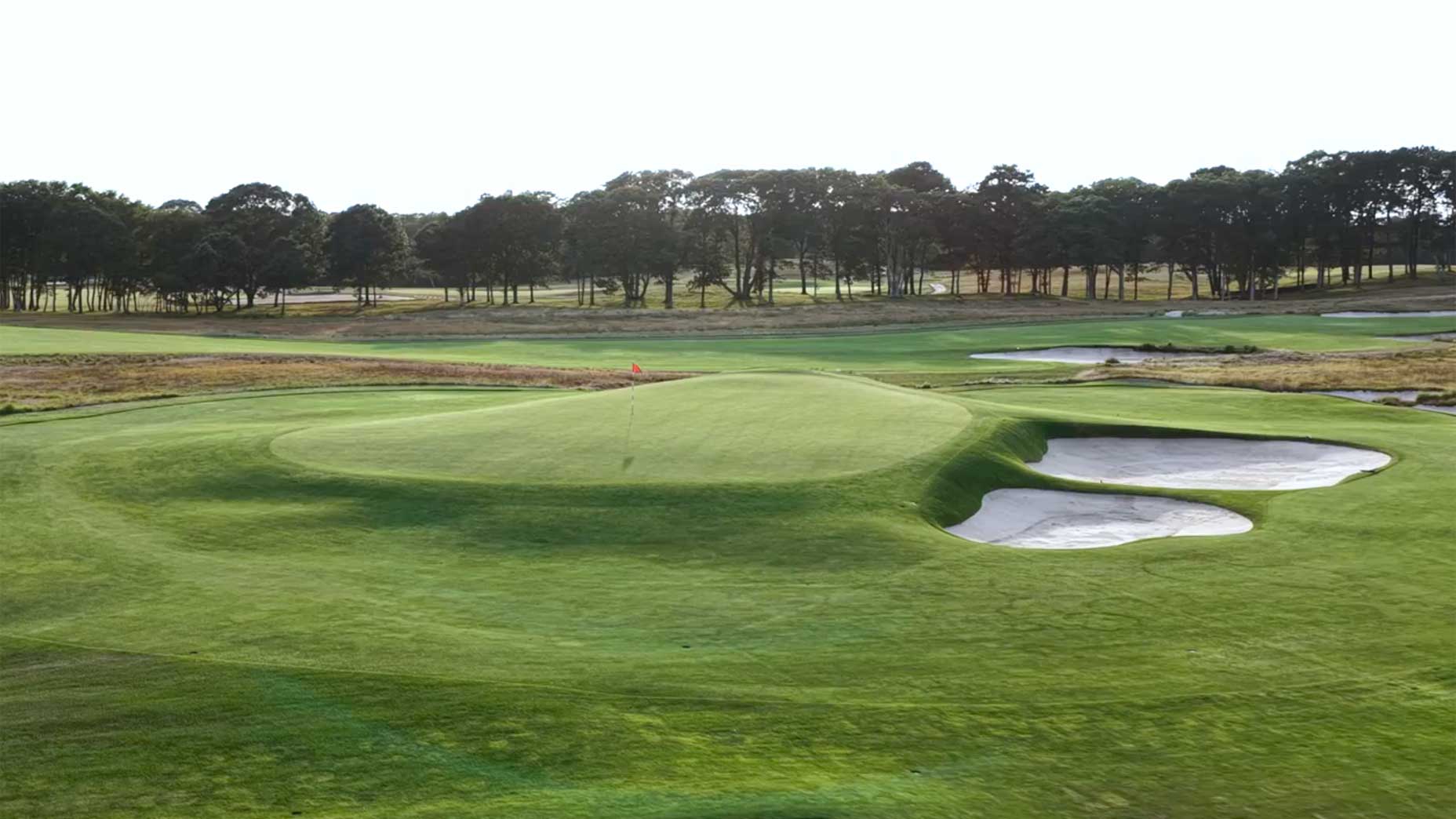 A well-manicured golf course with a green, several sand bunkers, a red flag, and trees in the background under a bright sky.