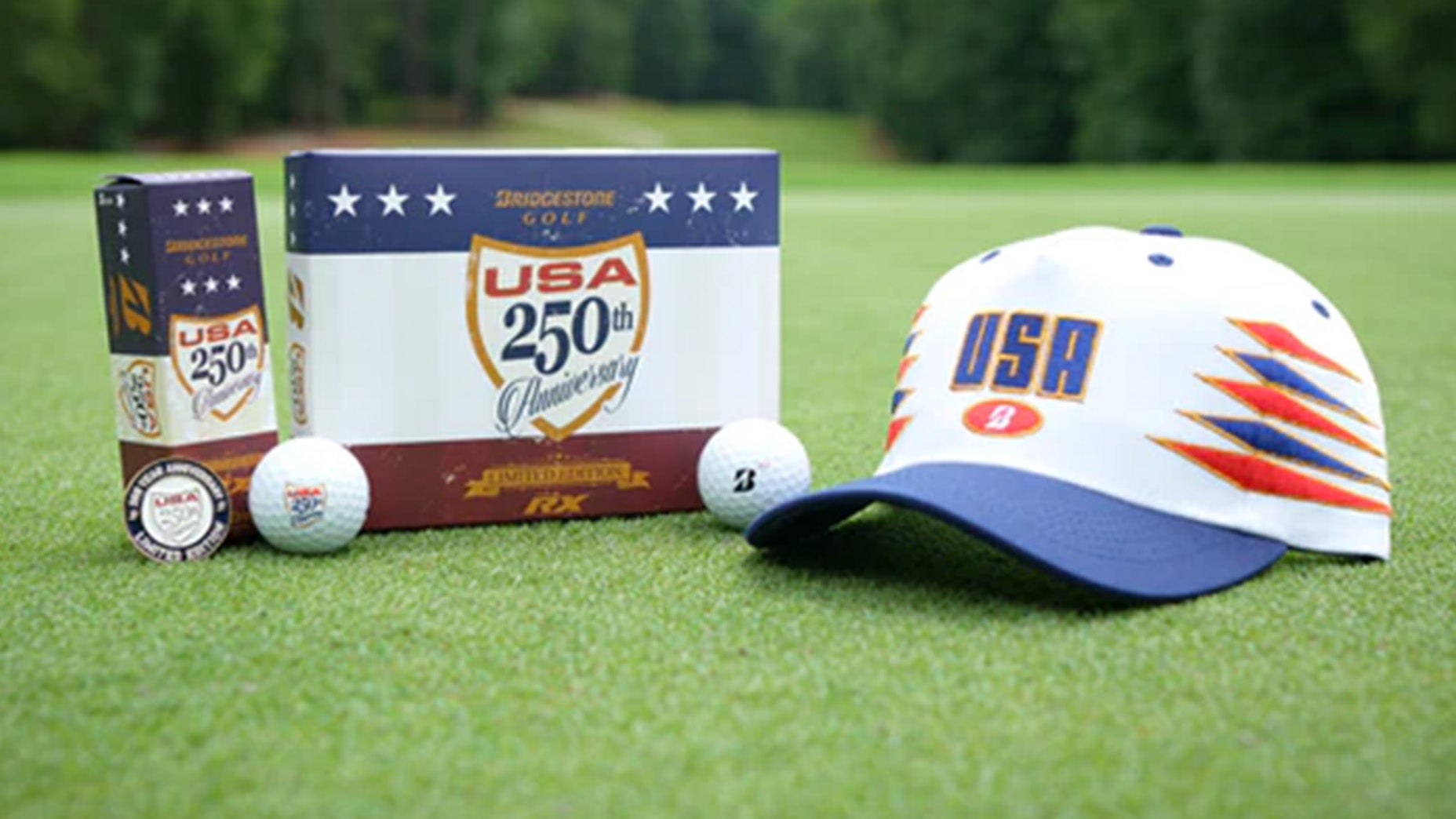 Golfausrüstung auf dem Grün: USA Bridgestone 250th Anniversary Golfballbox, rote, weiße und blaue Ausrüstung, darunter zwei Golfbälle, Ballmarker und weißer Hut mit blauem und orangefarbenem USA-Logo, mit verschwommenen Bäumen im Hintergrund.