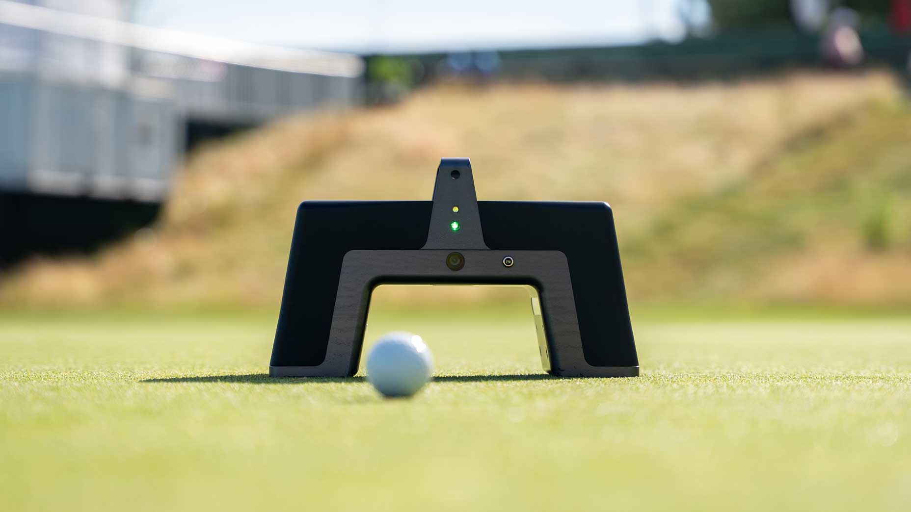 A close-up of a golf ball on a green positioned in front of a black golf training aid with green indicator lights, used for practice and alignment. Blurred grass and a structure are visible in the background.