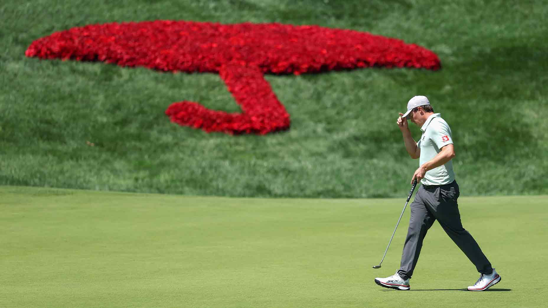 A golfer walks on a green, holding his putter and adjusting his cap, with a large red umbrella design made of flowers displayed on the slope behind him during the 2026 Travelers Championship Friday tee times.