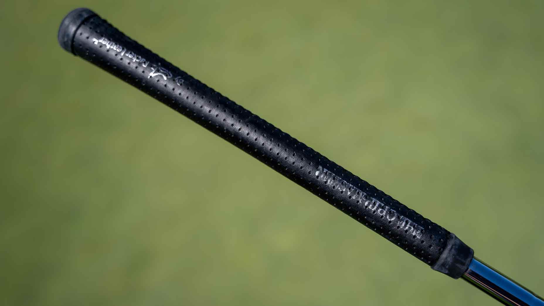 Close-up of a black, textured golf club grip featured in Bag Spy, with small dimples and white text, set against a blurred green background—just like something Min Woo Lee might have in his bag.