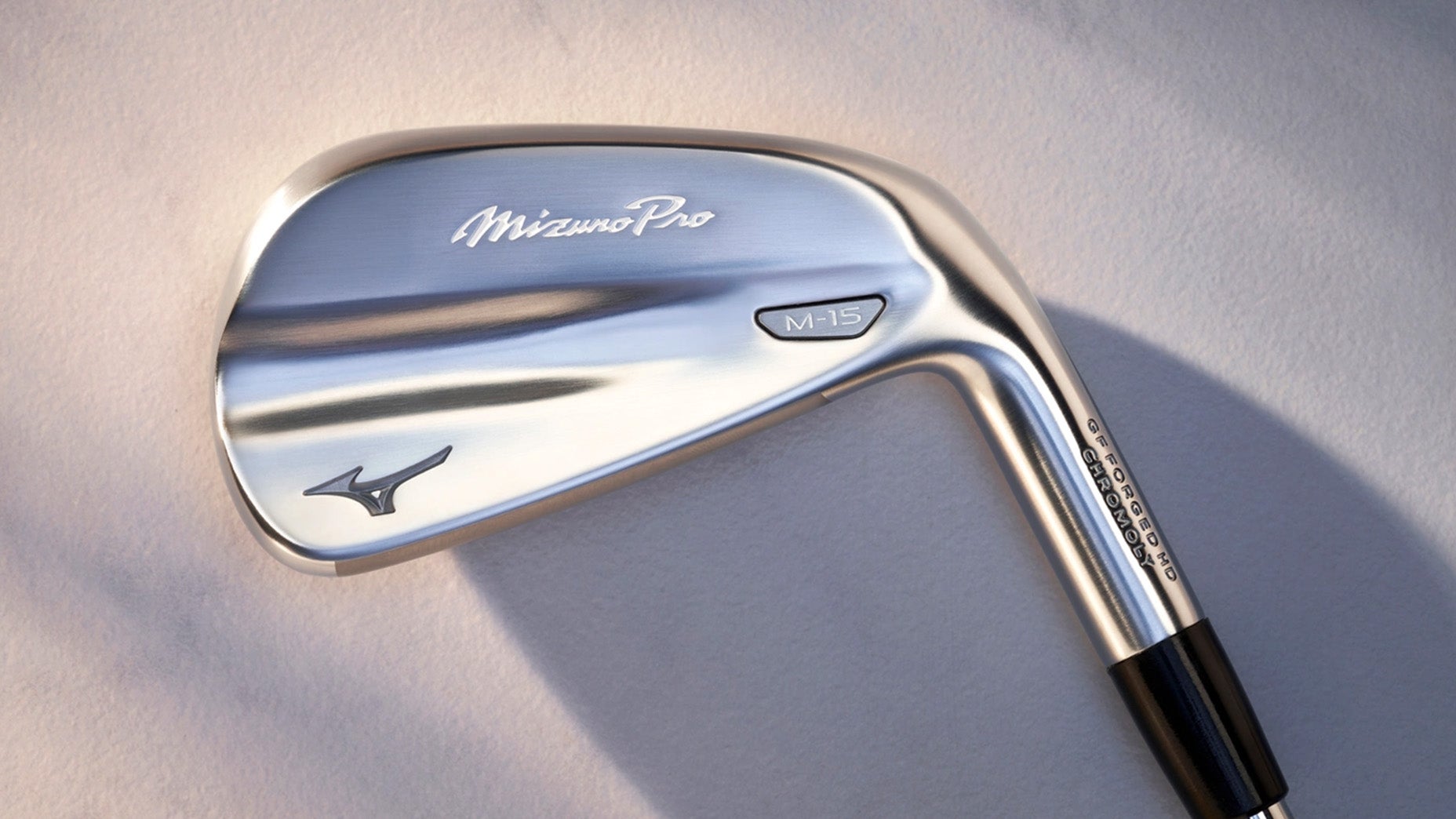 A close-up of a shiny silver Mizuno M-15 iron golf club head, showing the brand logo and model name engraved on the metal surface, with a soft light reflection highlighting its special features.