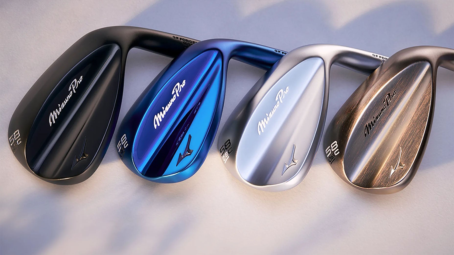 Four Mizuno Pro golf club wedges are displayed side by side in black, blue, silver, and bronze finishes, with their shiny surfaces reflecting light and the Mizuno logo and branding visible on each club.