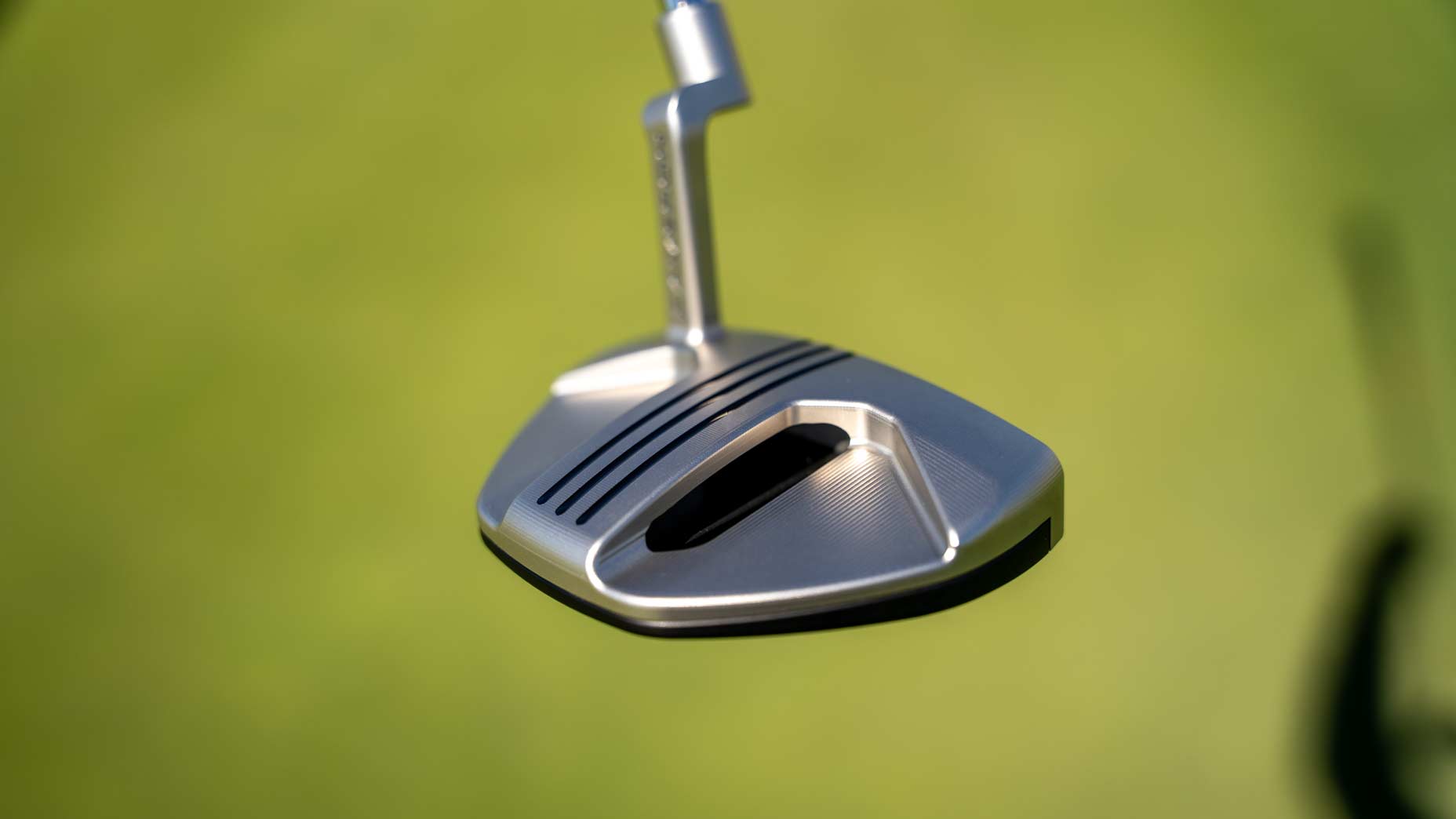 Close-up of a silver Scotty Cameron Phantom mallet putter with black grooves and a modern design, set against a blurred green background, likely a golf course.
