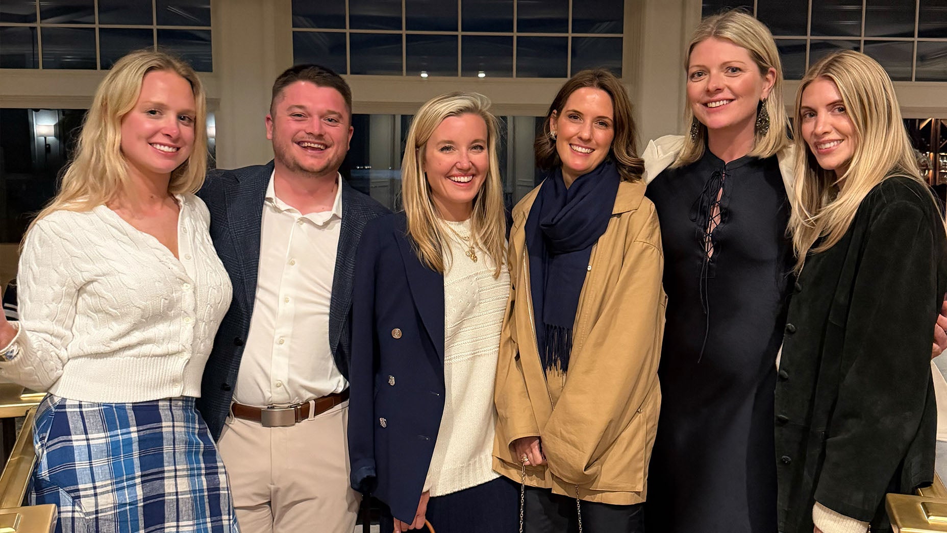Six adults, five women and one man, stand closely together indoors, smiling at the camera. Dressed in Ralph Lauren-inspired smart casual attire, they gather in a warmly lit room with large windows behind them.