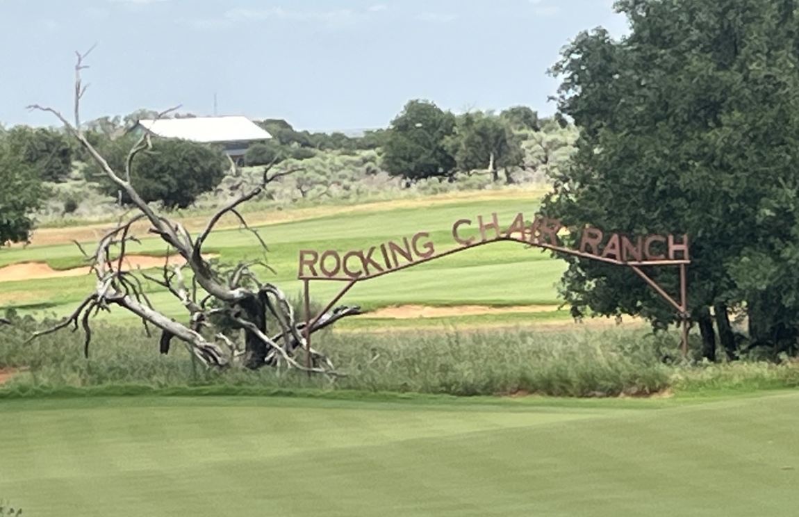 An arched metal sign reading ROCKING CHAIR RANCH stands at the edge of a green field, next to a leafless, twisted tree—hinting at classic cattle country—with trees and a white-roofed building in the background.