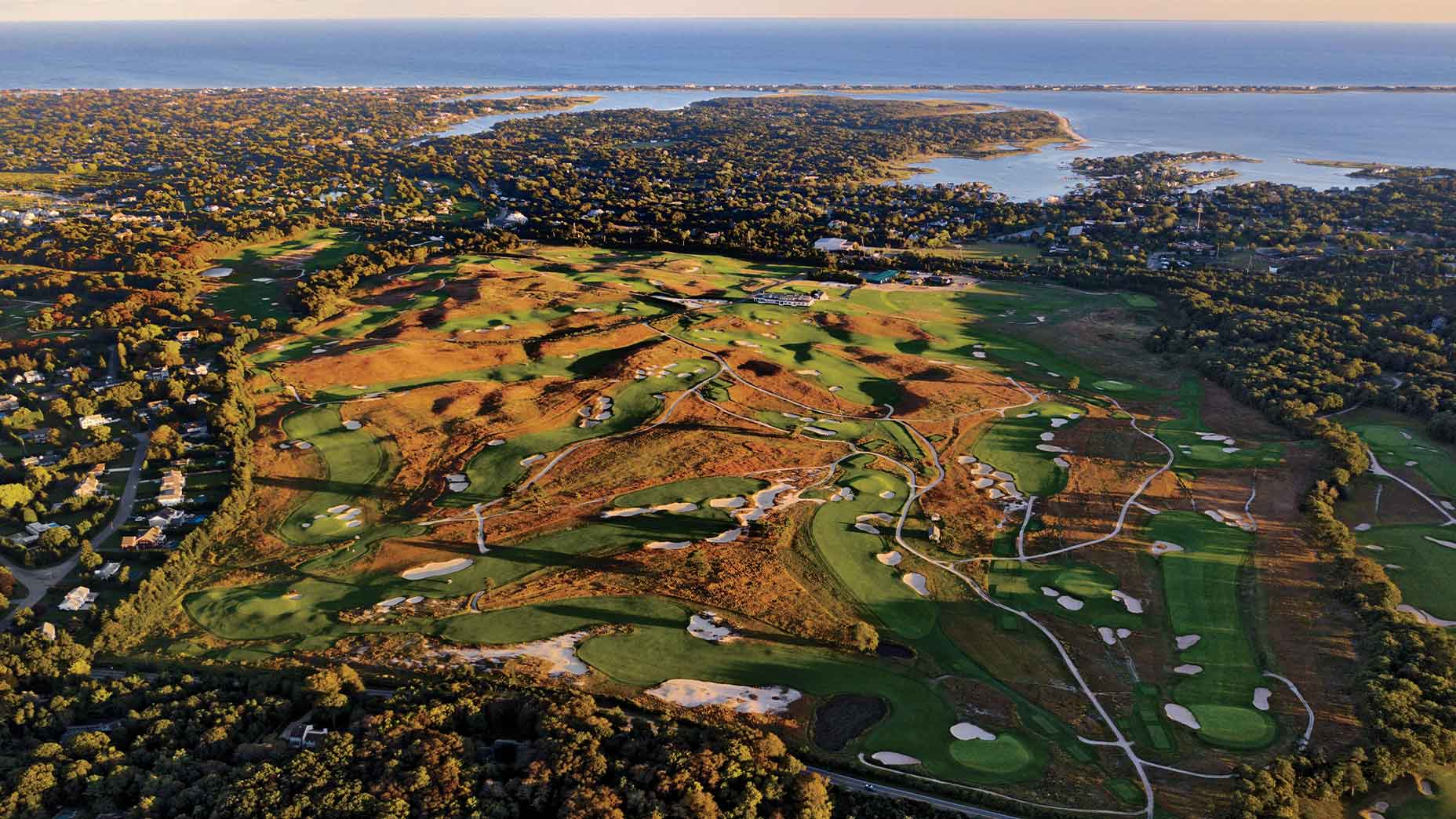 Aerial view of the expansive Shinnecock golf course surrounded by trees and residential areas, with water in the distance under a clear sky at sunset.