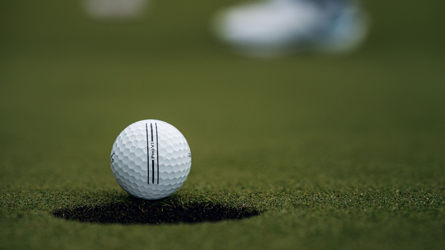 Close-up of a golf ball resting on the edge of a hole on a putting green, with blurred grass and a persons shoe in the background.