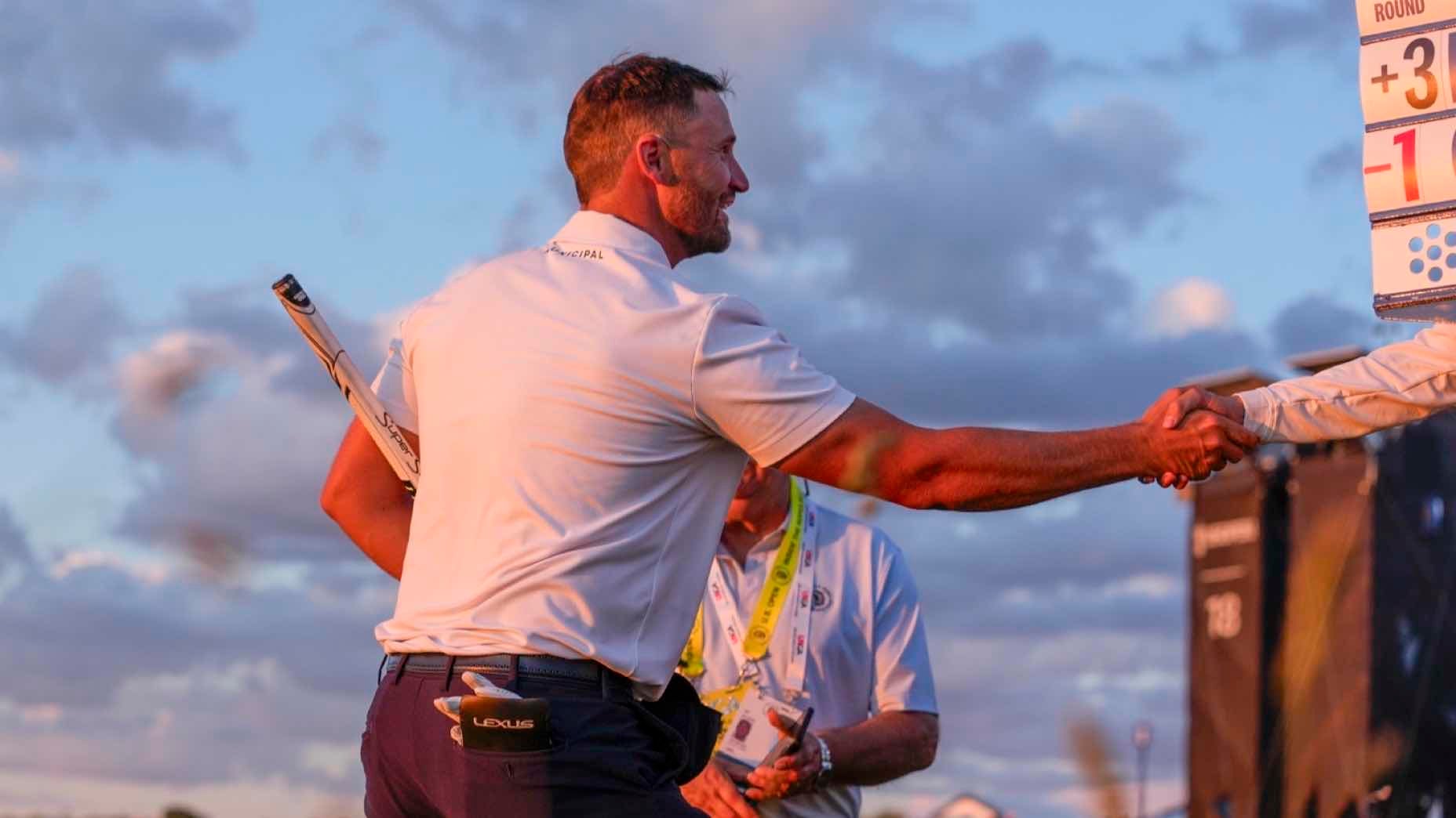 A golfer in a white shirt and navy pants shakes hands with someone off-frame, holding a golf club under his arm. Another person with a lanyard stands behind him, and a scoreboard is visible in the background against a cloudy sky.