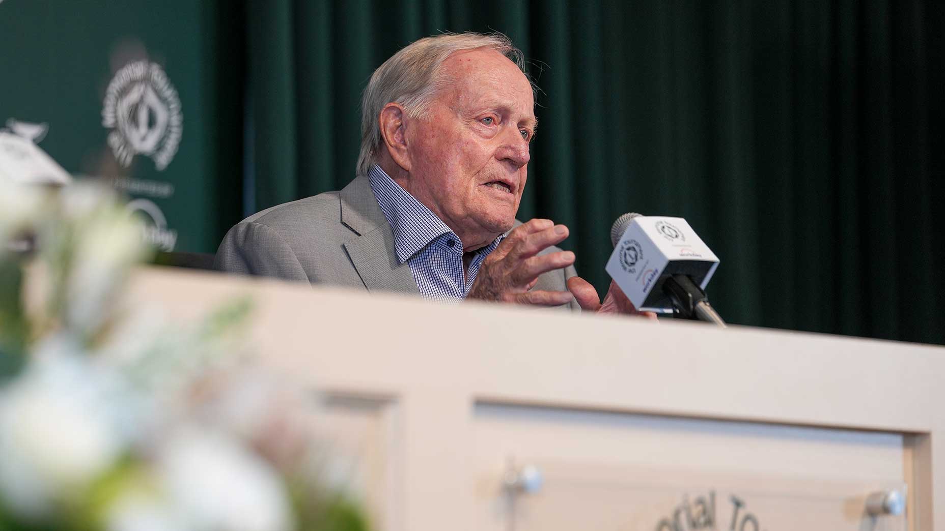 An older man in a suit speaks at a podium with microphones during a press conference, addressing the golf-ball rollback. Green curtains and a golf logo are visible in the background.