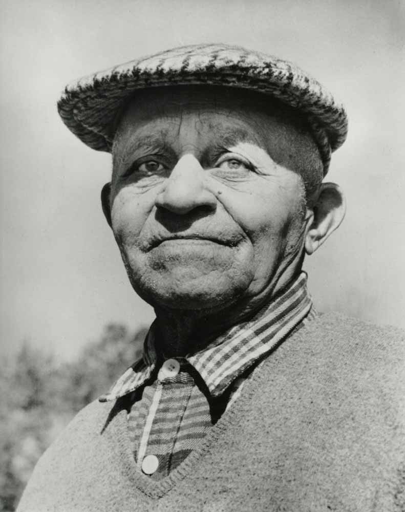 Black and white photo of an older man in a flat cap, plaid shirt, and sweater, smiling gently. He gazes slightly to the side; faint trees and sky blend in the blurred background. The portrait evokes memories of Shinnecock Hills at the 1896 U.S. Open.