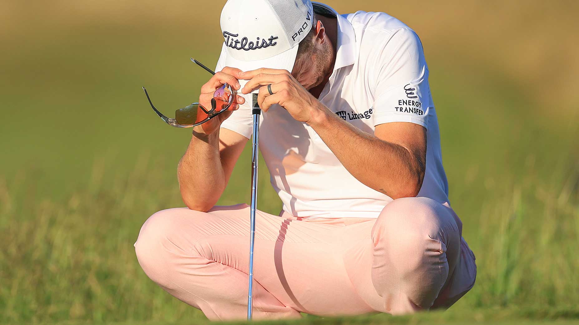 A golfer in a white cap and pink pants squats on the grass at Shinnecock Hills, holding sunglasses and resting his head on his hands, which are placed on top of a golf club during the U.S. Open.