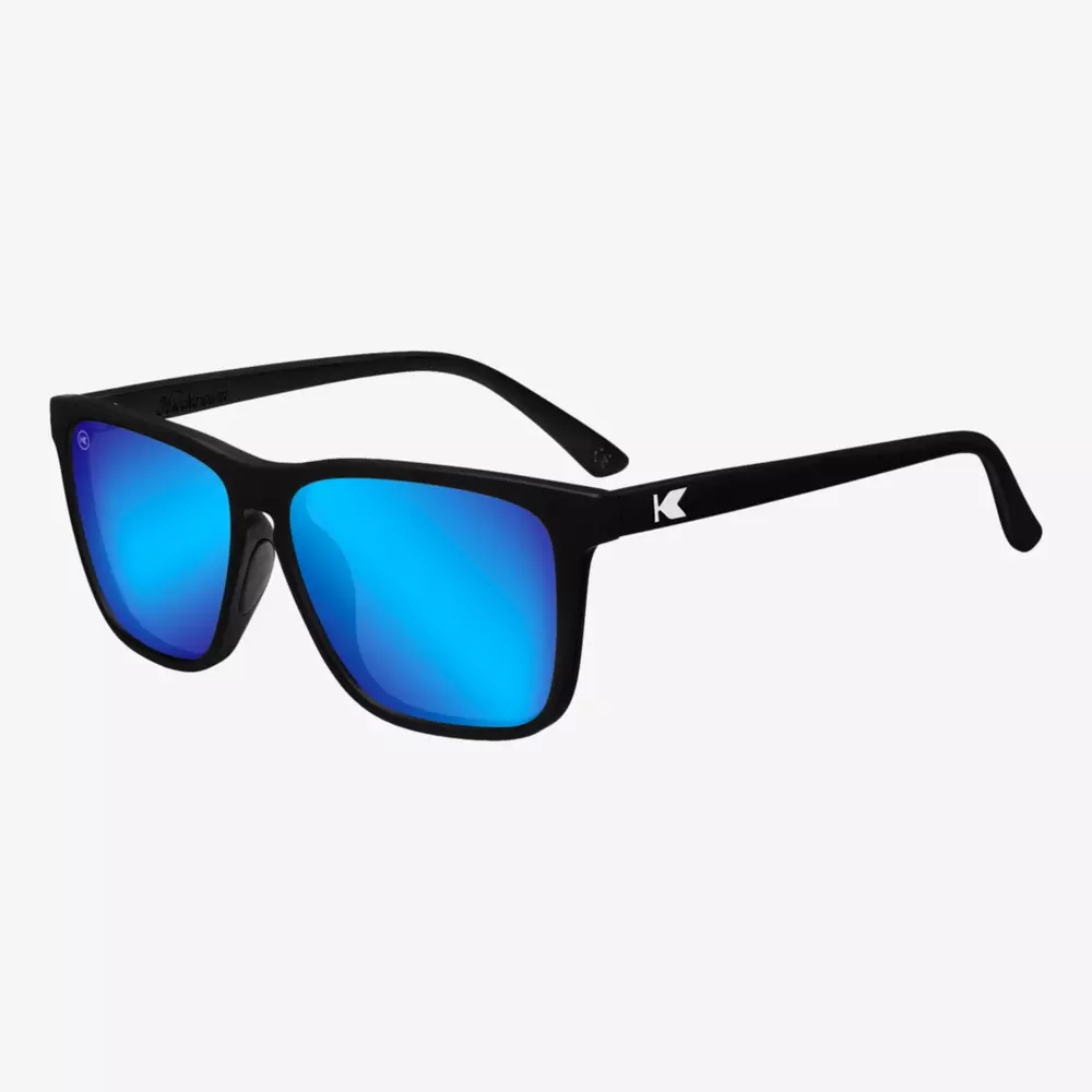 Schwarze Sonnenbrille mit leuchtend blauen verspiegelten Gläsern und einem kleinen weißen K-Logo an den Bügeln vor einem schlichten weißen Hintergrund.