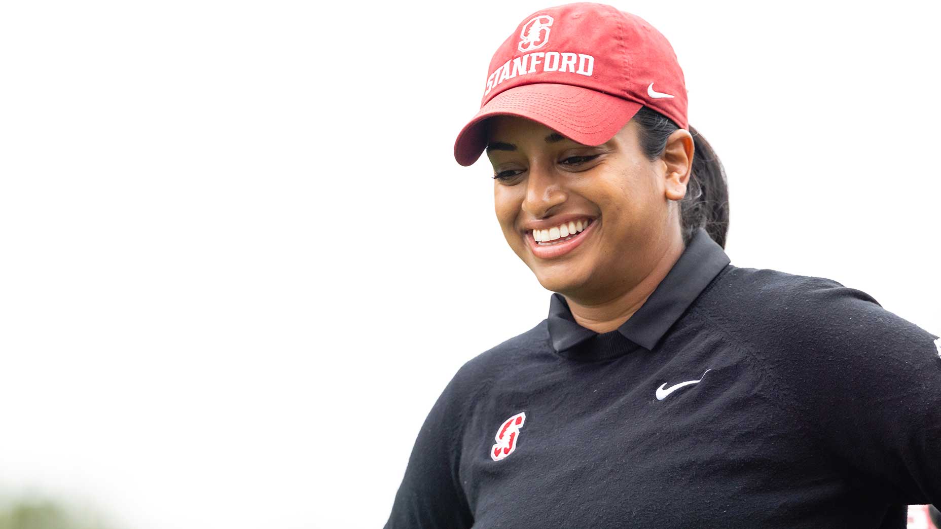 A woman, embodying her pro journey, wears a red Stanford cap and a black shirt with Stanford and Nike logos. Megha Ganne smiles while looking down, standing against a bright white background.