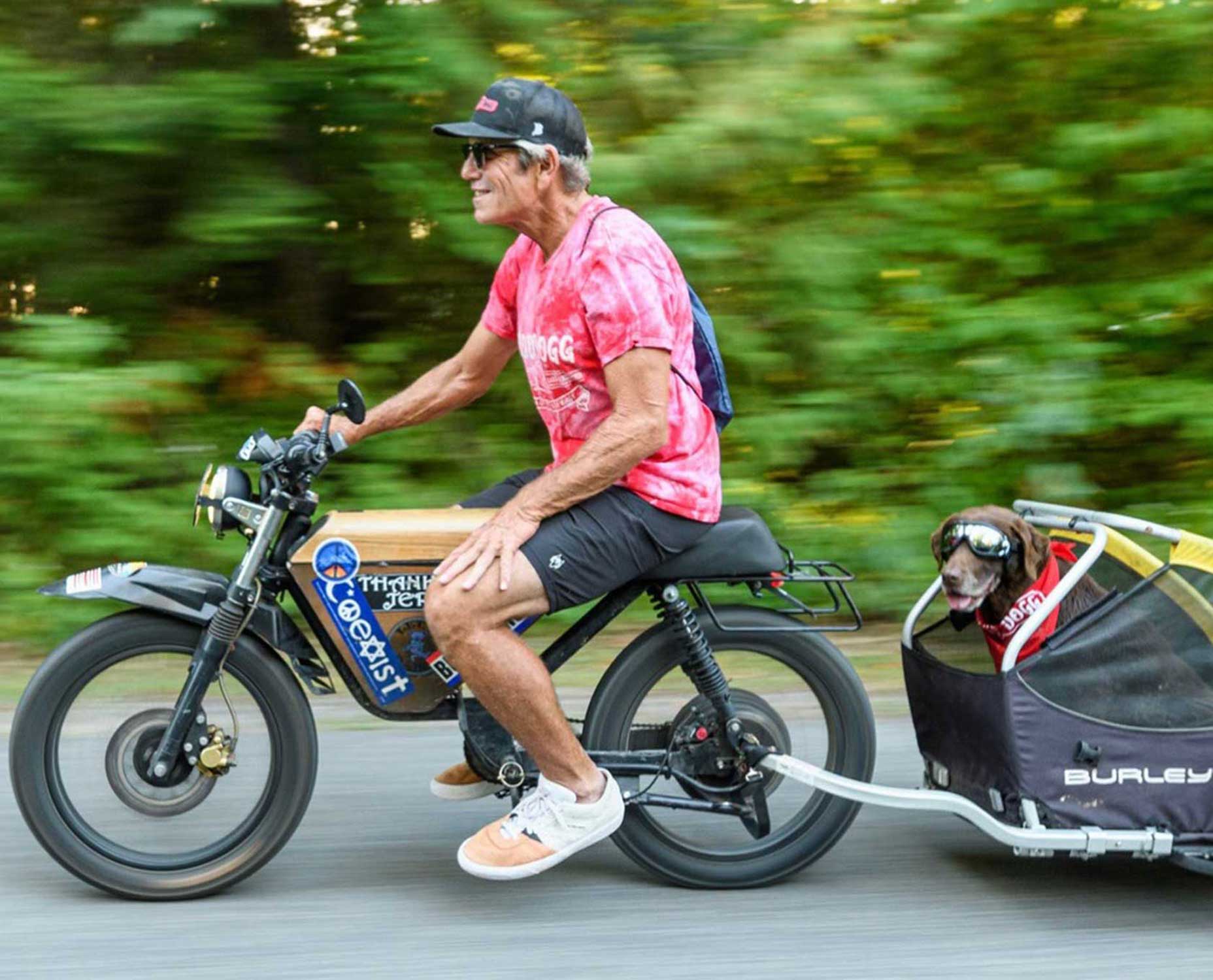 An older man wearing sunglasses and a red tie-dye shirt rides an electric bike, pulling a small trailer with a dog inside&mdash;like the Wizard of Odd on wheels. The background is blurred, capturing the motion and speed.