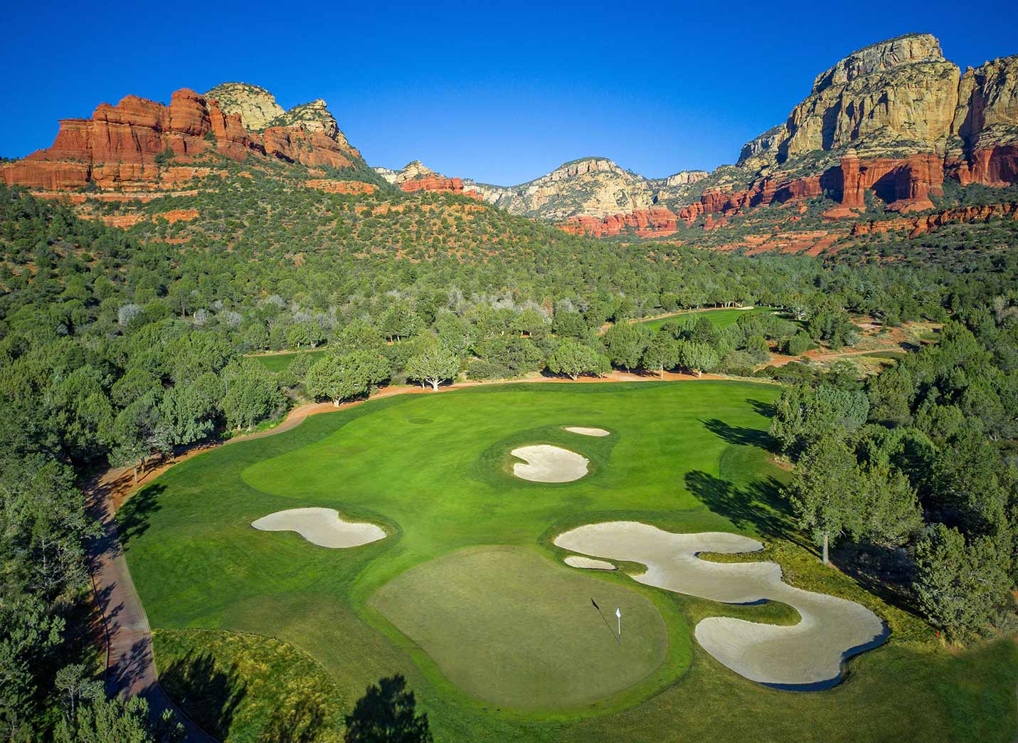 A green golf course with multiple sand bunkers is surrounded by trees and set against a backdrop of red rock mountains under a clear blue sky.