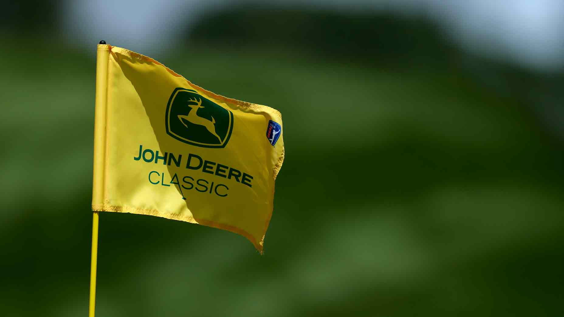 A yellow flag with the John Deere Classic logo featuring a deer, fluttering against a blurred green background during Round 4 TV coverage.