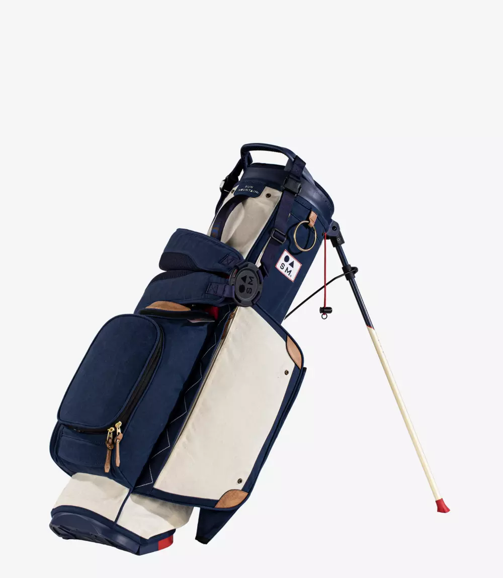 A navy blue and cream golf bag with brown accents stands upright on its fold-out legs. The bag features multiple zippered pockets and a padded strap for carrying.