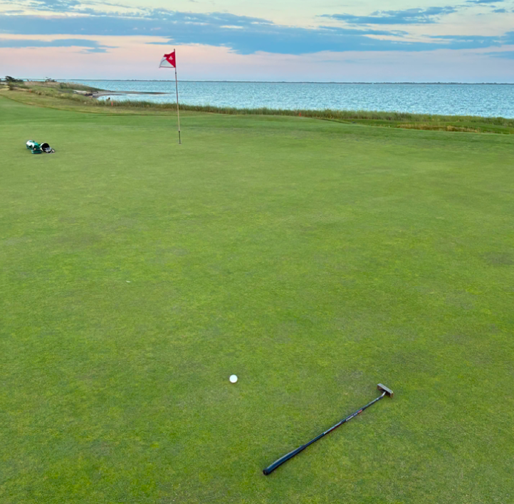 A golf ball and putter rest on a green near a red-flagged hole, with a golf bag in the distance and a large body of water under a partly cloudy sunset sky—evoking classic South Shore golf like Shinnecock or the U.S. Open.