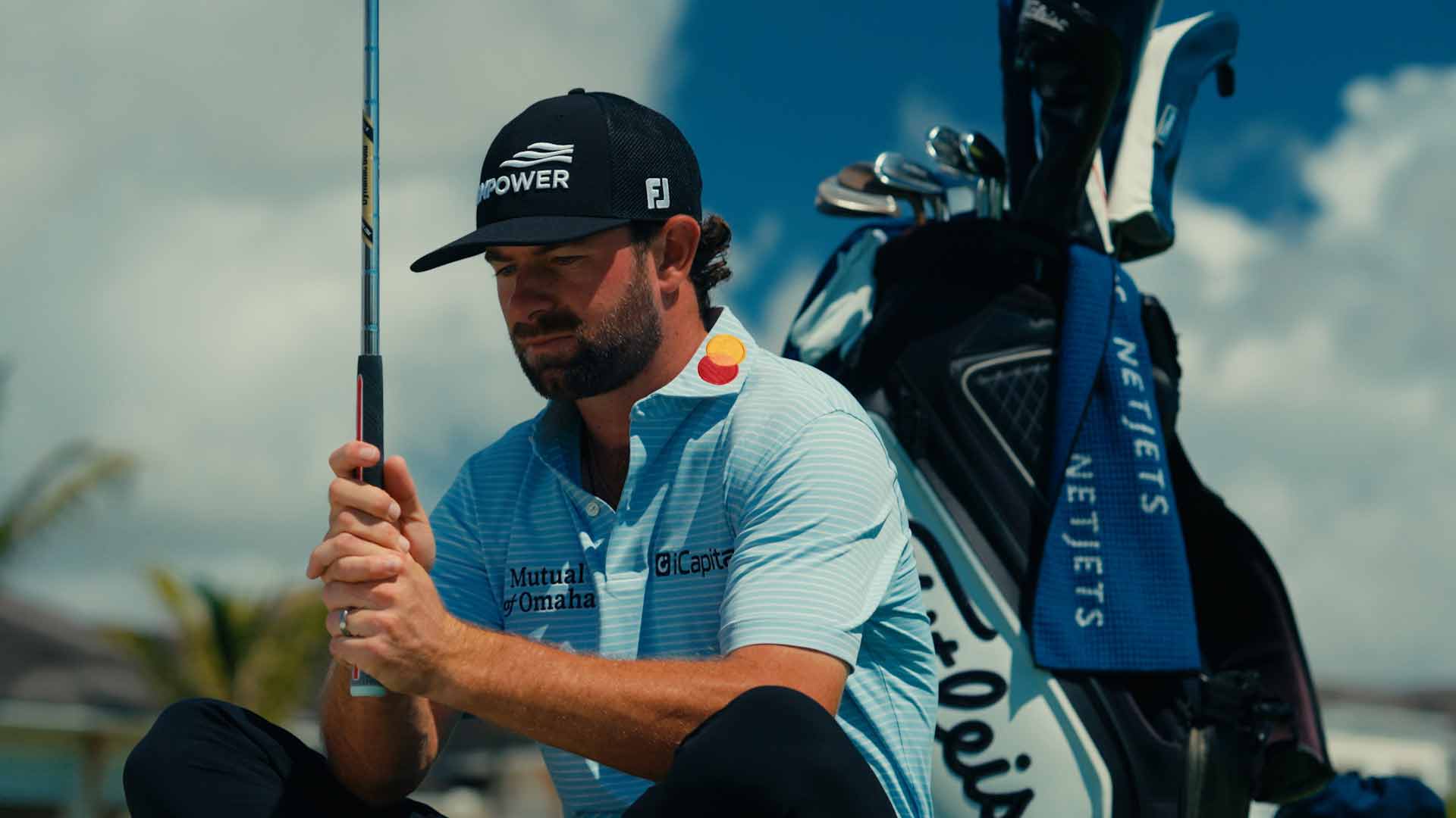 A male golfer wearing a cap and blue shirt squats on a golf course, holding a club. There’s a golf bag and towel beside him, with palm trees and blue sky in the background.