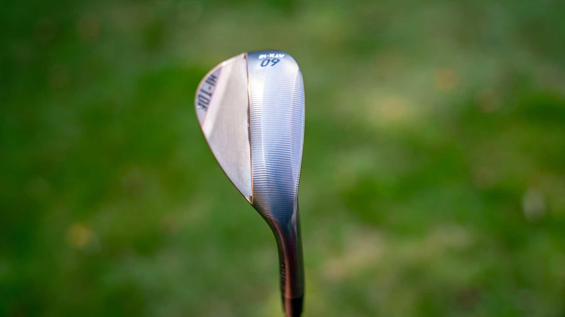 A close-up of a TaylorMade Hi-Toe wedge with 60 marked on the forged clubface, set against a blurred green background, likely grass.