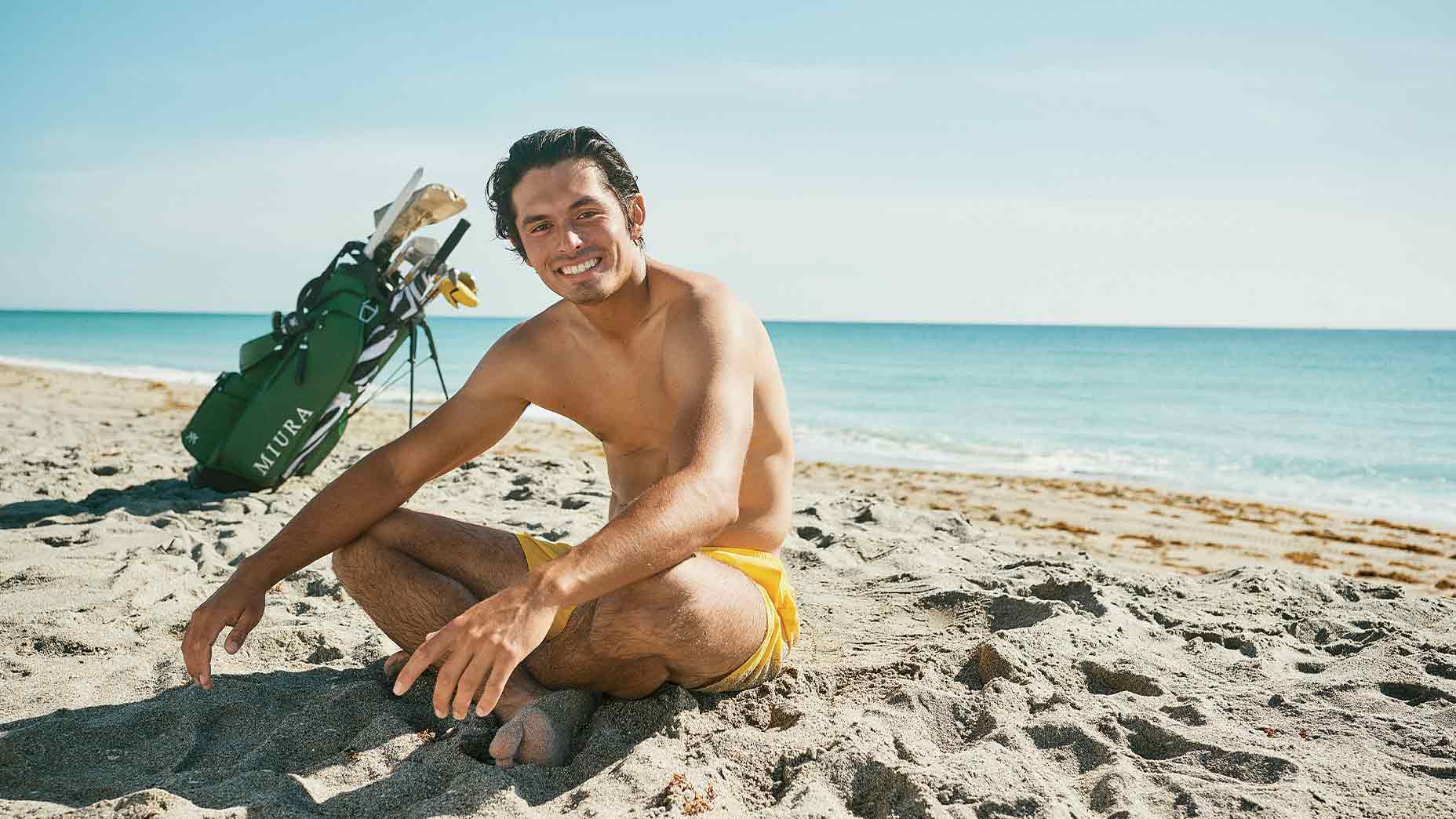 A man in yellow swim trunks sits smiling on a sandy beach near the ocean, with a green golf bag and clubs behind him under a clear blue sky—a scene reminiscent of Fat Perez, one of golf’s digital boom online creators, merging relaxation and the love of the game.