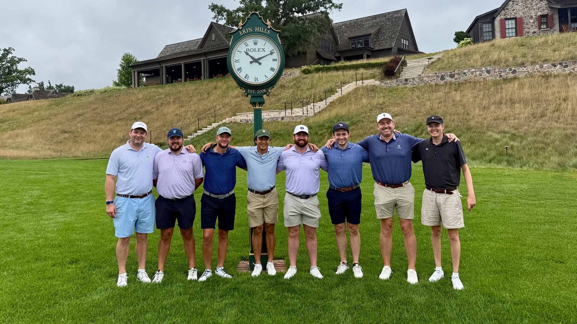 Eight men stand arm in arm in front of a large outdoor Rolex clock on a golf course, enjoying their Wisconsin golf trip, with clubhouses and grassy hills in the background.